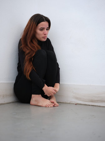 Low angle view of a sad young woman in the corner of a room dressed in black barefoot and on the...