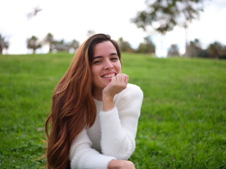 A young woman in the park happily smiling at the camera with her hand on her chin.の写真素材