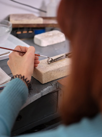 a woman working in her artisan jewelry workshop using a small brush on a ring to perform her work on...
