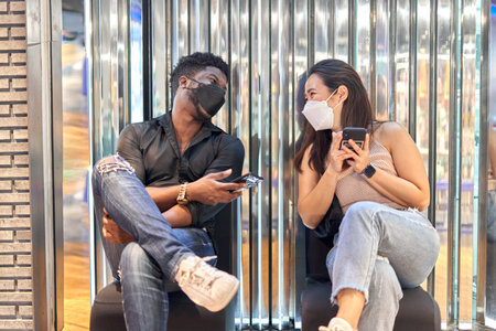 Multiethnic friends wearing mask using the mobile and sitting on a shopping mallの写真素材