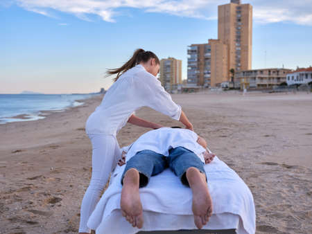 A young chiromassage therapist giving a massage to young adult man to treat his back injury on a beach in Valencia with buildings on background.の写真素材