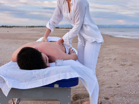 A young man lying on a massage table receiving a chiromassage treatment on the beach in Valencia by an unrecognizable young chiromassage therapist.の写真素材