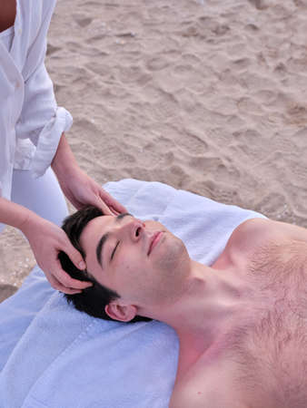 A relaxed young man lying on a massage table receiving a head massage on a beach in Valencia by a young chiromassage therapist.の写真素材