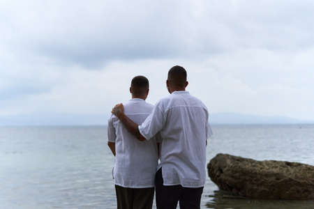 Gay couple gazing at the horizon of the ocean as they embrace in the eveningの写真素材