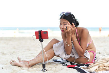 Asian young female streamer applying make-up for a live broadcast from a beachの写真素材