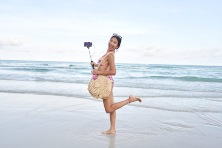 Asian streamer posing funny and holding a selfie stick with a mobile on a beachの写真素材