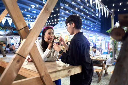 Non binary person feeding ice cream to her partner at urban night fair