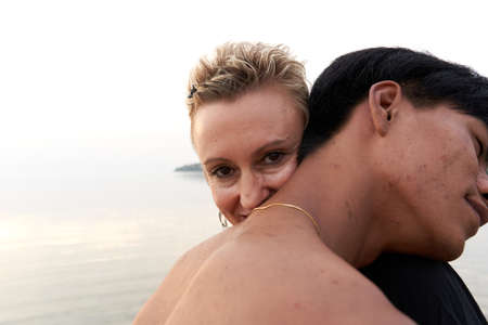 Caucasian woman smiling while hugging a young asian man next to the sea