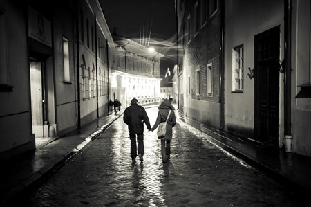 A young couple holding hands walking at night in the old town street, cobbled street wet from the rain, autumn eveningのeditorial素材