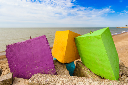 Three different colours - purple, yellow and green concrete breakwater blocks on the beach, in the backgroundblue cloudless sky, yellow sand and coastline の写真素材