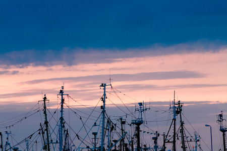 Plenty of masts of fishing boats and sailing boats in sunset background, visible ropes, rigging,...