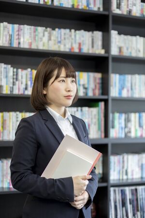 Woman holding a book and notebook in front of a bookshelfの写真素材