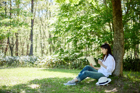 Image of early summer of a Japan woman in her 30s relaxing and reading in the forestの写真素材