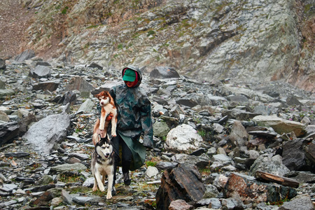 Girl in cloak with hood carries puppy of  red Siberian husky. Mountainous terrain, rocky landscape, rain.の写真素材