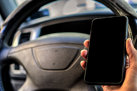 Cell phone with blank screen in hand on car steering wheel background. Black screen of phone for...