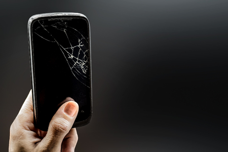 Hand is holding a cell phone with broken glass against a dark background. Broken smartphone with cracking glass of the phone black screen.の写真素材