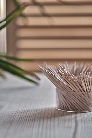 Wooden toothpicks stand on wooden table. On background of window and green twig tropical plant. Side view, vertical photo. Copy space.の写真素材