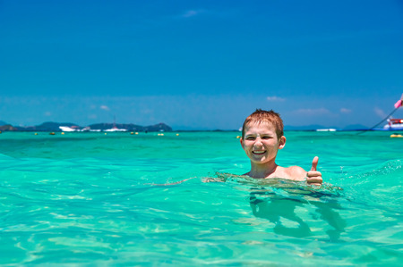 Boy (10 years old) bathing in turquoise water tropical sea. Child is smiling while showing thumb up. Seascape with bright blue sky and shimmering clear sea. Background, copy space.の写真素材