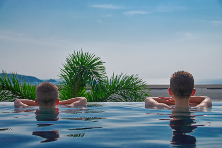 Two children in outdoor pool with his hands on board look at ocean. Rear view. Copy space. For...