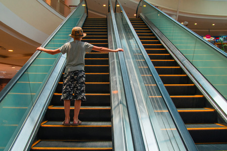 Boy rises up on an escalator in shopping center. Child in hat, t-shirt and breeches. Rear view. Copy spaceの写真素材