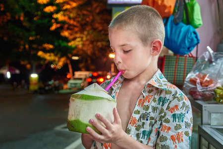 Cute boy drinks coconut milk from the opened green fruit of coconut through a cocktail tube. Child on the night street quenches thirst.の写真素材