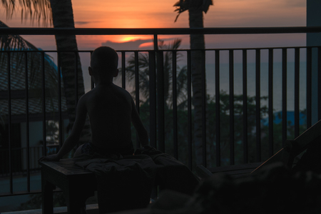 Silhouette of a child on a tropical sunset over the sea. Boy sits on the balcony and watching the sunset.の写真素材