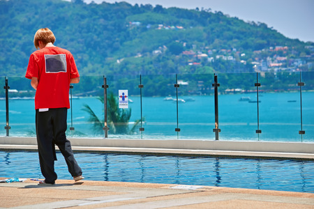 Back view portrait young man standing by the outdoor pool and using smart phone. Guy in red tshirt....