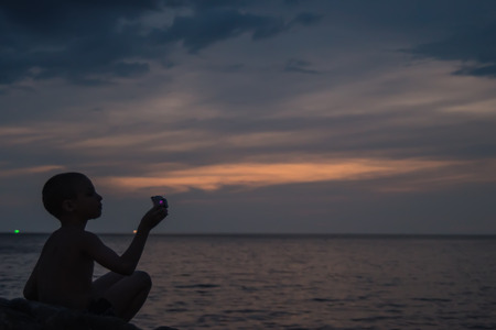 Side view portrait of a happy boy relaxing sitting on the rock and watching the sunset sea in the background. Baby holding an action camera. Copy space.の写真素材