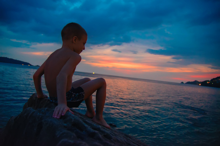 Side view portrait of a happy boy relaxing sitting on the rock and watching the sunset sea in the background. Copy space.の写真素材