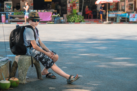 Concept traveling with Infant or children. Boy age 12 in a hat, sunglasses, shorts, tshirt and with backpack is sitting on the street. Child on summer vacation, adventure. Copy space.の写真素材