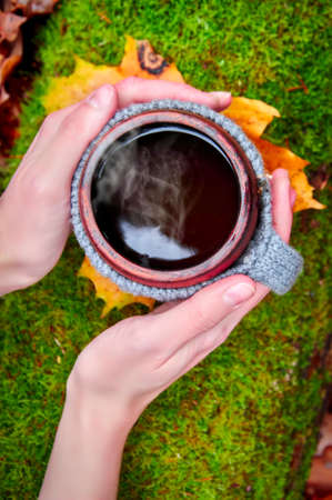 A mug of hot tea coffee in women's hands. View from above against the background of green moss. Picnic, hike.の写真素材