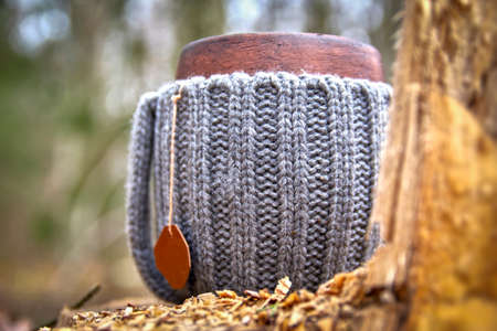 Clay mug in woolen knitted cover. Steam from the mug on the background of autumn forest and sunlight, close up.の写真素材