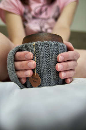 Cozy morning in bed with glanny mug of coffee, womens hands hold a mug in knitted cover. Close-up.の写真素材