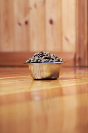 Metal bowl with dry pet food stands on the wooden floor.