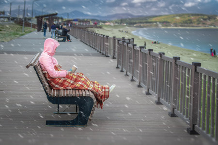 Girl dressed in jacket, wrapped in blanket sits with cup coffee on the bench, shore cold gloomy seaの写真素材