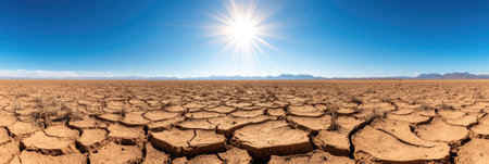 A vast arid terrain with dry, cracked ground stretches under a blazing sun, framed by distant mountains.の素材