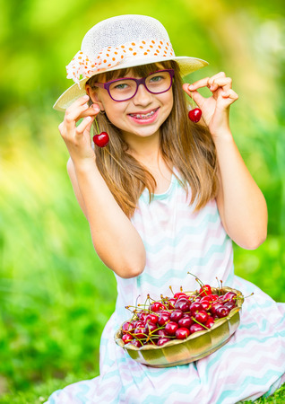 Child with cherries. Little girl with fresh cherries. Young cute caucasian blond girl wearing teeth braces and glasses. Portrait of a smiling young girl with bowl full of fresh cherries.の写真素材