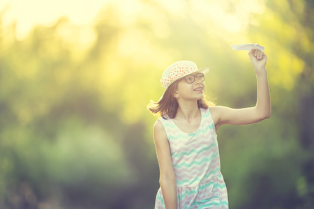 Cheerful pre-teen girl playing with paper plane on the park at sunrise. Girl with glasses and teeth braces.の写真素材