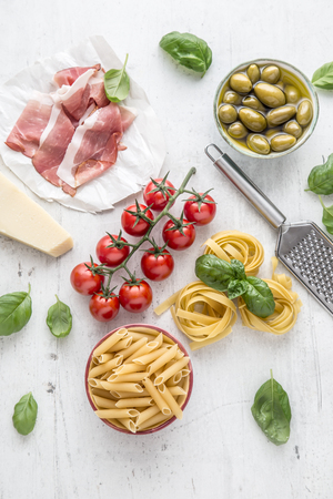 Italian or mediterranean food cuisine and ingredients on white concrete table. Tagliatelle pene pasta  olives olive oil tomatoes parmesan cheese prosciutto and basil leaves on white concrete board.の写真素材