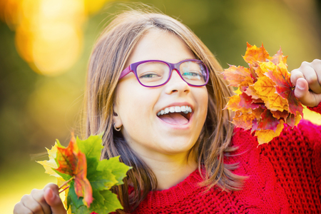 Happy fall girl smiling and joyful holding autumn leaves. Beautiful young girl with maple leaves in red cardigan.の写真素材