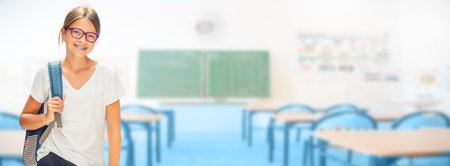 Portrait of modern happy teen school girl with bag backpackand class room as background. Girl with...