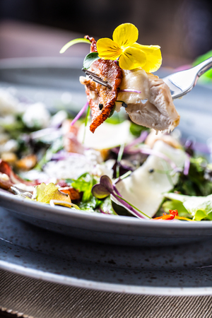 Close-up pieces of chicken breast baked bacon and flowers decoration on fork. In the background plate vith mixed salad.の写真素材