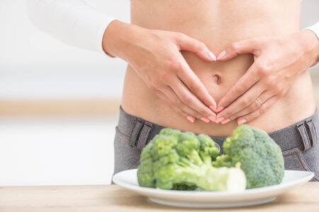 Woman with sports figure on her belly shows heart shape. Fresh broccoli in plate on kitchen table.の写真素材