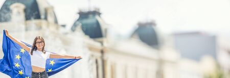 Cute happy young girl with the flag of the European Union in front of a historic building somewhere in europe.の写真素材