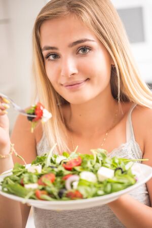 Young happy blonde girl eating healthy salad from arugula spinach tomatoes olives onion and olive oil.の写真素材