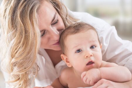 Mother and baby boy son playing on a white bed. Mothers tenderness and kisses of a toddler child.の写真素材