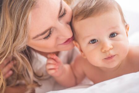 Mother and baby boy son playing on a white bed. Mothers tenderness and kisses of a toddler child.