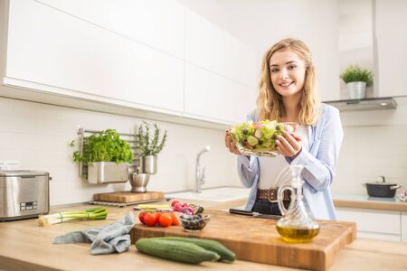 Young happy blonde girl preparing healthy salad in home kitchen.の写真素材
