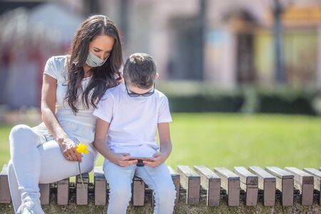 Young woman holding a flower sits next to a boy in the town, both looking into a cell phone in his hands and wearing face masks.の写真素材
