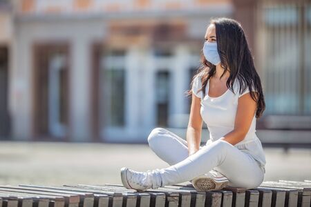 Good looking girls sits alone on the bench in the city center wearing a face mask during Covid-19 pandemic.の写真素材